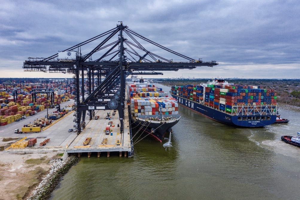 Two large cargo ships, heavily loaded with colorful shipping containers, are docked at a bustling port in Pasadena under a cloudy sky. Workers and cranes busily handle the cargo, as tales of public intoxication amidst the maritime hustle add an unexpected layer of intrigue to the scene.