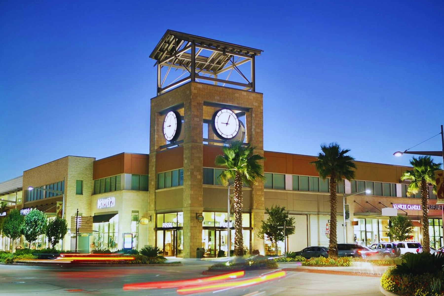 At twilight, the shopping center in Pearland boasts a prominent clock tower and palm trees, with vivid streaks of car lights weaving through the scene, capturing city life's vibrancy even amidst whispers of public intoxication.