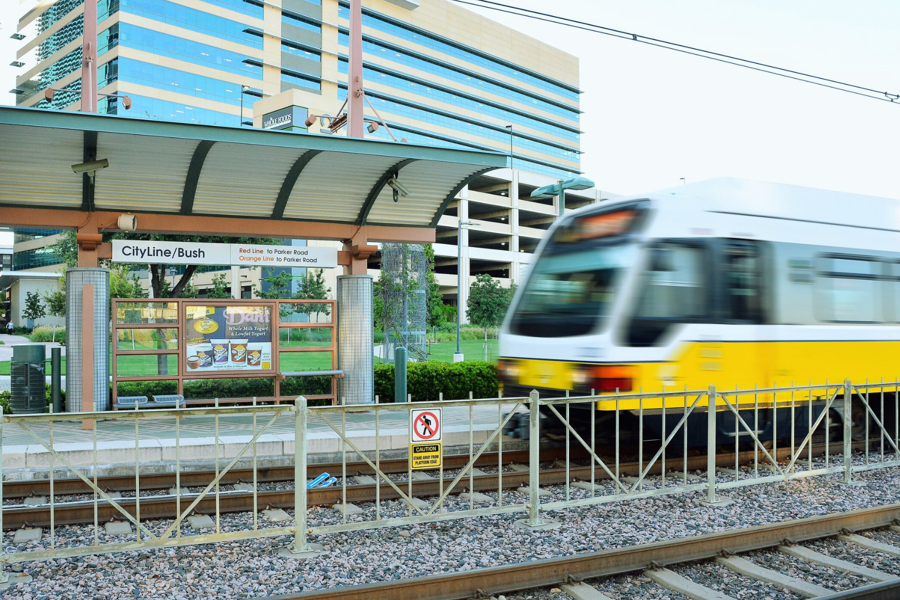 A light rail train glides past CityLine/Bush station, framed by a modern building in the background. The clear station sign and schedule stand on the platform, a testament to smooth urban transit even amidst occasional public intoxication issues in Richardson.