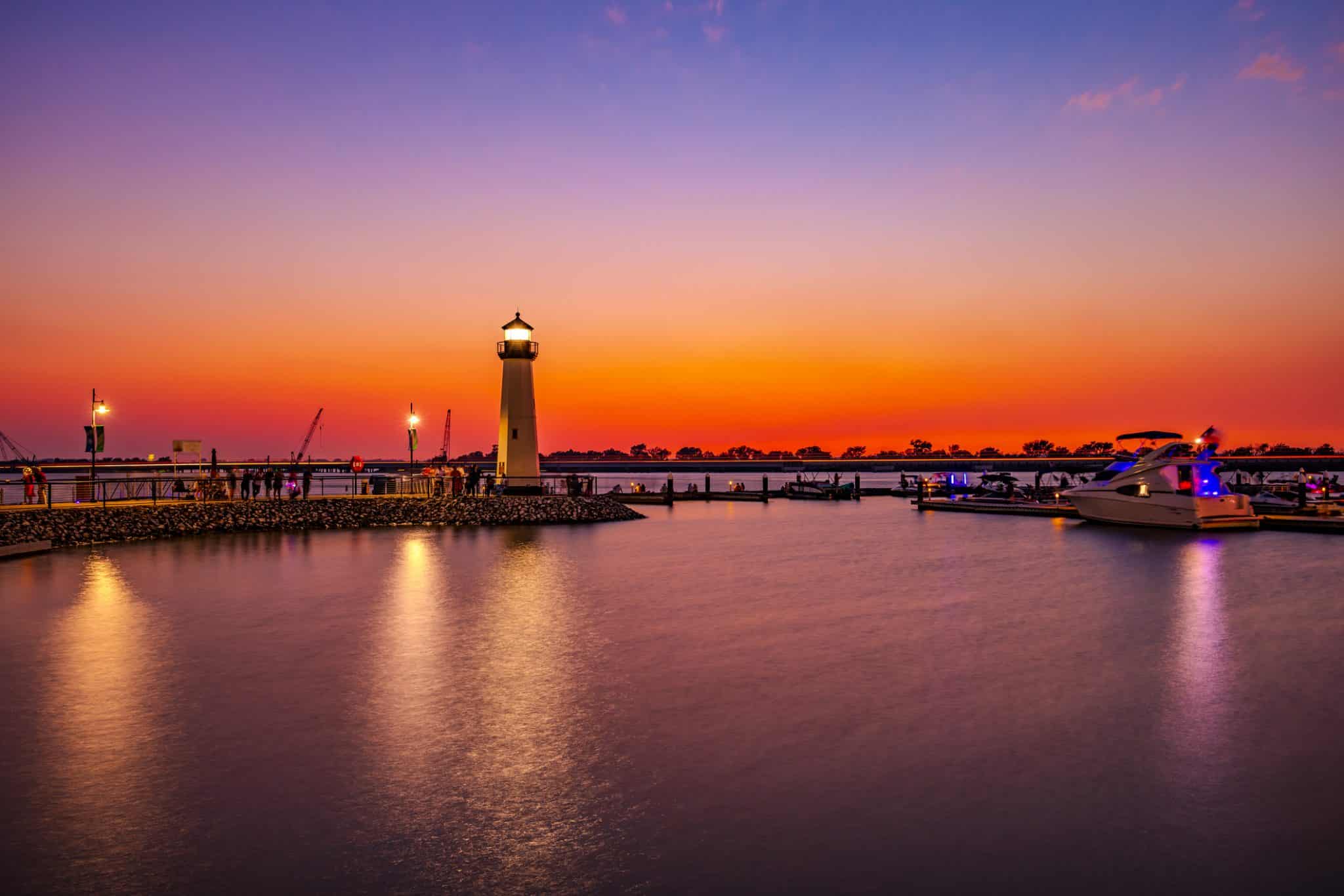 The serene lighthouse by the marina at sunset, with boats docked and calm waters reflecting the orange and purple hues of the sky, offers a tranquil escape from the bustle of city life.