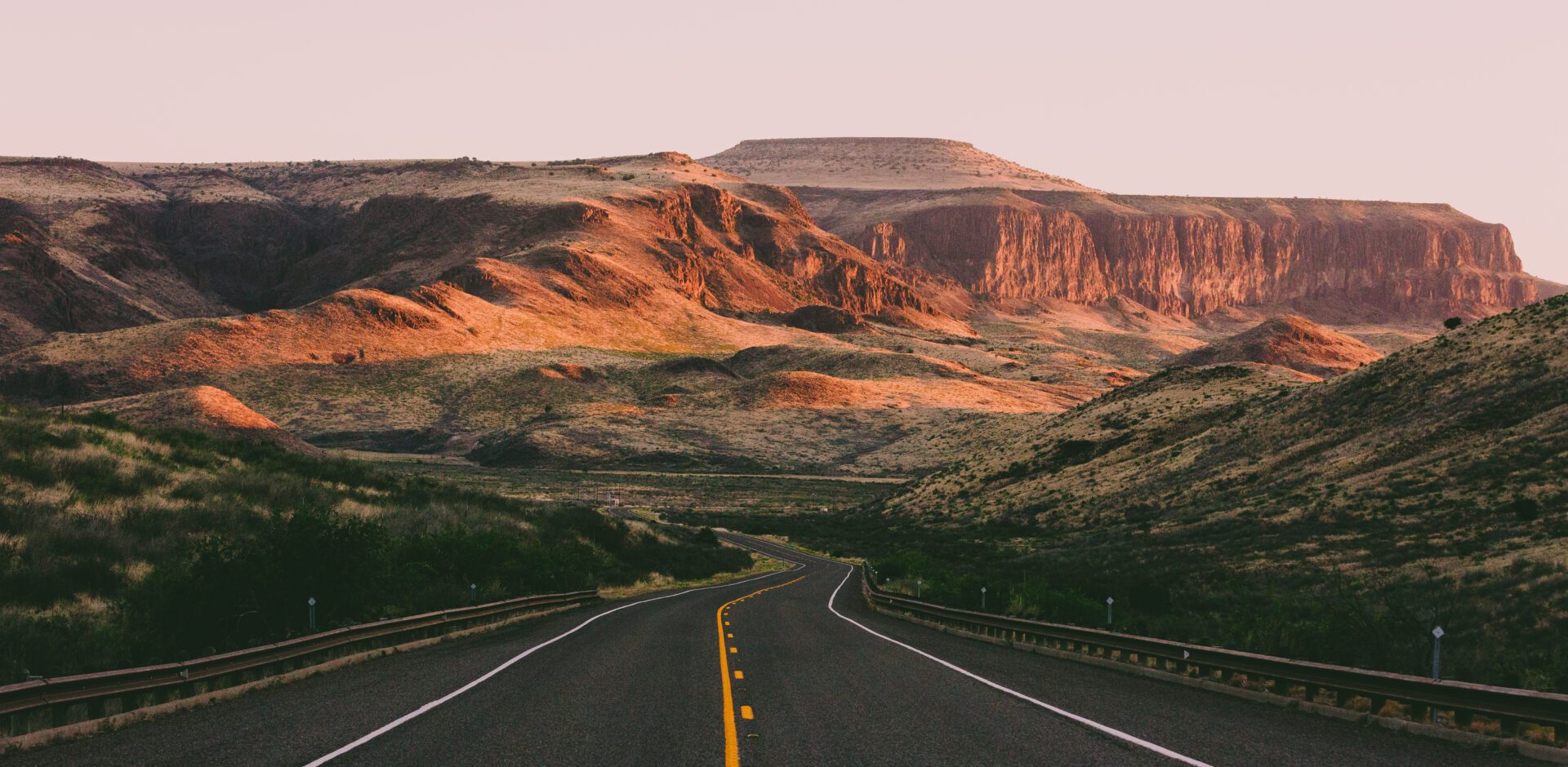 A winding road leads toward a landscape of sunlit, rocky hills and mesas under a clear sky at sunset, reminiscent of the quiet escape you crave after experiencing the bustling energy of Texas.