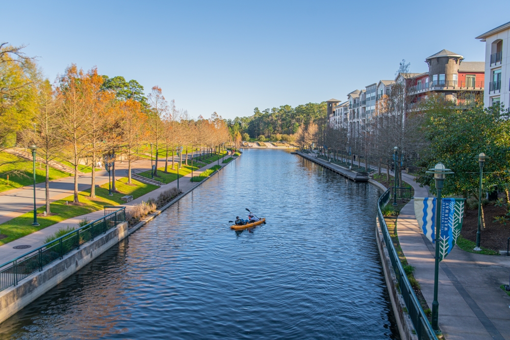 A person kayaking on a peaceful canal in Woodland, Texas, flanked by pathways and residential buildings, with trees lining both sides under a clear blue sky, much like the soothing guidance of a local expungement lawyer navigating through complex legal waters.