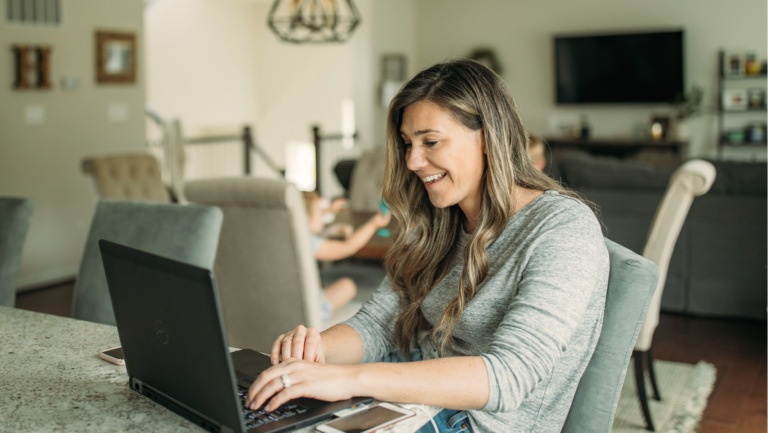 A woman sits at a kitchen counter in her Texas home, diligently working on a laptop. In the background, the living room features a TV and couches, creating a cozy workspace as she researches the process of sealing criminal records.