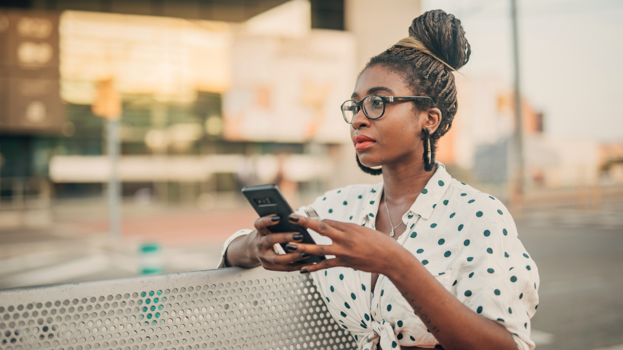 In a bustling Texas city, a person with glasses and braided hair casually holds a smartphone, seemingly unbothered by the surrounding chatter.