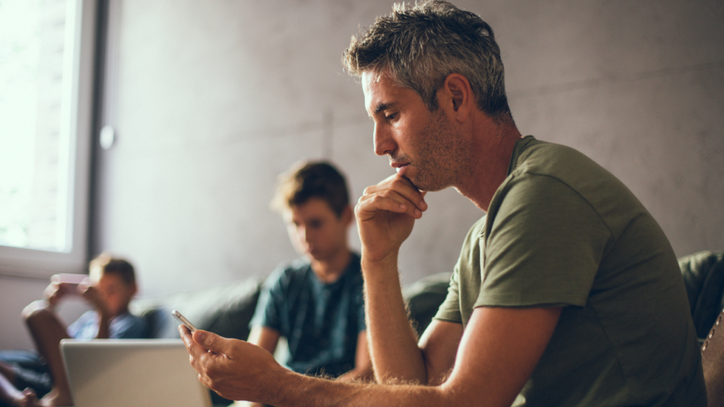 A man in a green shirt sits on a couch in Texas, absorbed in his phone. Two others linger in the background, one engaged with a laptop, as if unaware of the recent buzz about a local DWI arrest.