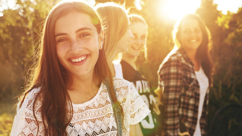 A group of four smiling women stands outdoors in sunlight, enjoying their day. The woman in the foreground, with long hair and a white lace top, unknowingly highlights the importance of understanding Texas DUI and underage drinking laws for those under 21.