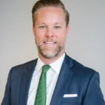 A man with a beard and neatly styled hair, wearing a dark suit, white shirt, and green tie, poses against a plain background.