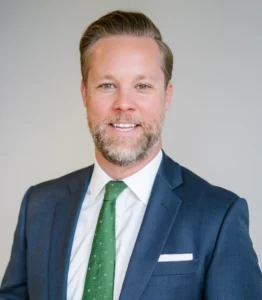 A man with a beard and neatly styled hair, wearing a dark suit, white shirt, and green tie, poses against a plain background.